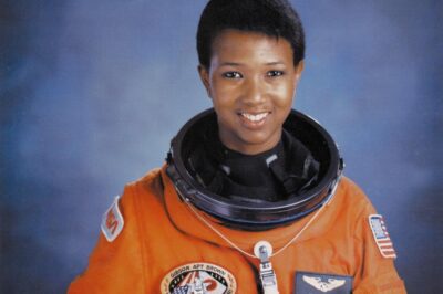 A woman astronaut in an orange spacesuit against a blue background.