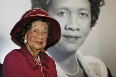 Elderly woman in a red hat with a large black-and-white portrait behind her.
