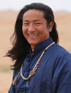 Smiling man with long hair wearing a blue traditional outfit and necklace.