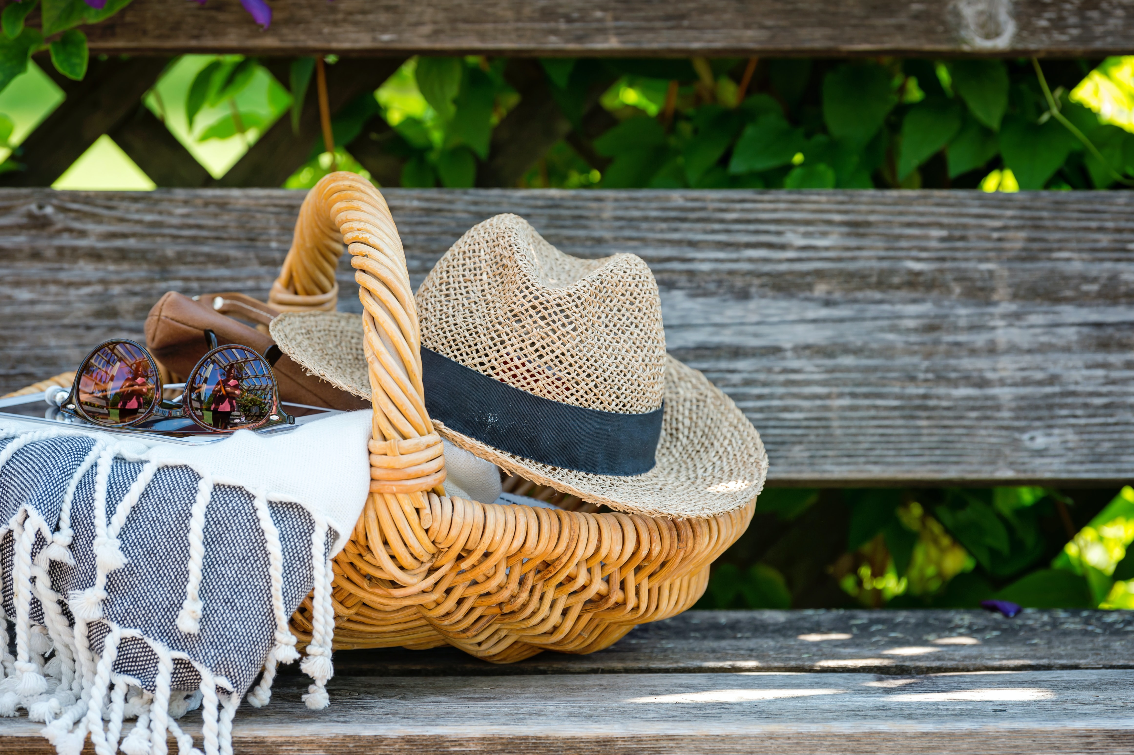 A wicker picnic basket with a straw hat and sunglasses on a rustic wooden bench.