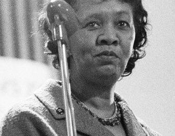 Black and white photo of a woman speaking at a microphone.
