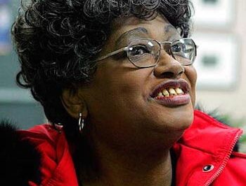 Smiling elderly woman with curly hair and glasses in a red jacket.