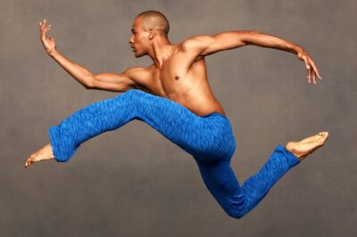 A man in blue pants leaps energetically against a neutral background.