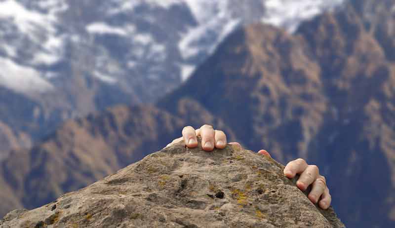 Hands gripping a rock during a mountain climb with snowy peaks in the background.