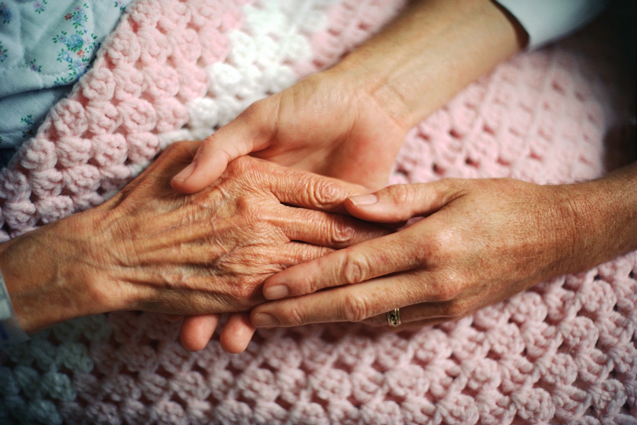 Two elderly hands gently holding each other on a knitted blanket.